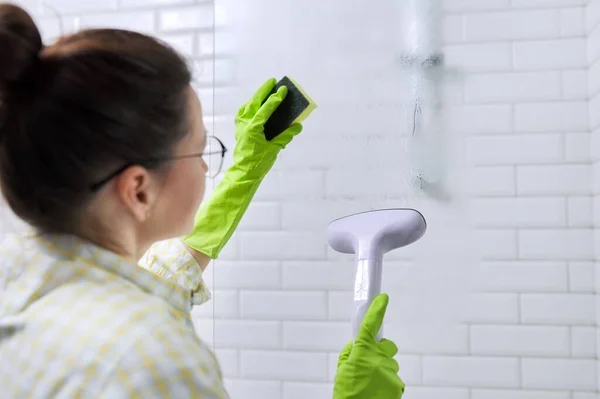 Woman housewife doing house cleaning in bathroom, female washing glass with shower steam. Using steam cleaner for quick cleaning