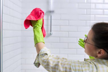 Kadın banyoda temizlik yapıyor, evde. Dişi temizlikçi, duş camını mikrofiberli bir bez ile parlatıyor..