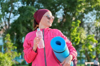 Spor rüzgarlığı giymiş, yoga minderli su şişesiyle dışarıda yürüyen olgun bir kadın. Orta yaşlı insanlarda aktif sağlıklı yaşam tarzı, spor, fitness