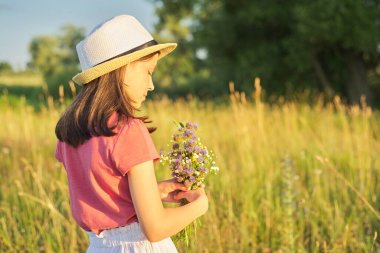 Şapkalı güzel romantik kız çocuğu, güneşli çayırlarda bir buket kır çiçeği toplayan elbise, resimli manzara, altın saat, fotokopi alanı. Çocukluk, yaz, doğa, güzellik, çocuk konsepti.