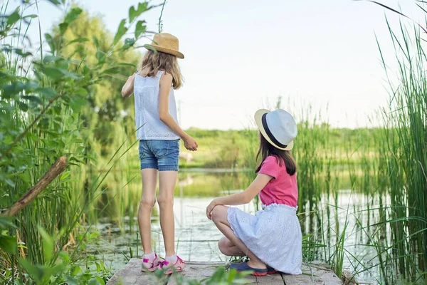 两个漂亮的女孩坐在芦苇丛中的湖边木码头上 回头看 大自然 快乐的童年 乡村风格 — 图库照片