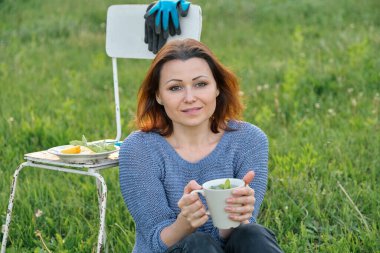 Bahçedeki çimenlerde oturan, gün batımında dinlenen, naneli çay içen orta yaşlı güzel bir kadın. Hobiler, doğa, bahçe işleri, altın saat