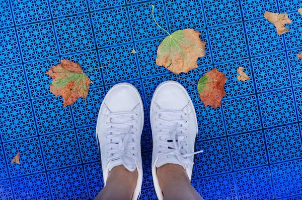 Autumn in city, close-up of legs in sneakers and fallen first yellow leaves, blue surface of the playground background