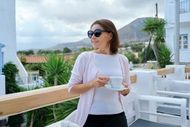 Açık hava restoran tatil köyünde elinde bir fincan kahveyle dolaşan olgun, güzel bir kadın. Güzel resimli dağ manzarası. Eğlence, eğlence, yaz, hafta sonu, turizm, orta yaşlı insanlar