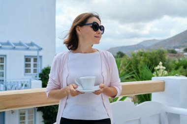 Açık hava restoran tatil köyünde elinde bir fincan kahveyle dolaşan olgun, güzel bir kadın. Güzel resimli dağ manzarası. Eğlence, eğlence, yaz, hafta sonu, turizm, orta yaşlı insanlar