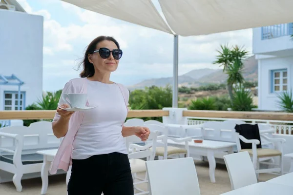 Mature beautiful woman walking with cup of coffee in outdoor restaurant resort hotel. Beautiful picturesque mountain landscape. Recreation, leisure, summer, weekend, tourism, travel middle-aged people