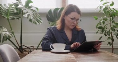 Pijamalı güzel olgun bir kadın dijital tablette haber okuyor.