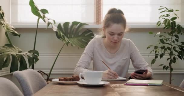 Chica adolescente sentada en casa en la mesa escribiendo lecciones usando un teléfono inteligente para aprender — Vídeos de Stock