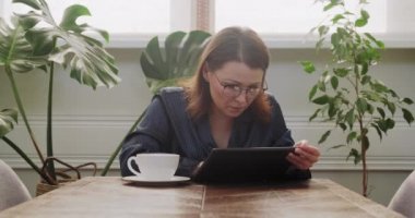 Elinde çay fincanı olan pijamalı olgun bir kadın dijital tablet okuyor, kötü haber.