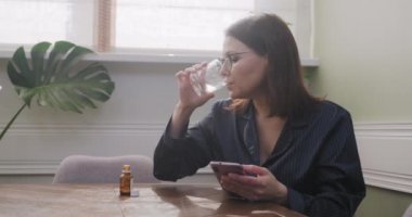 Heyecanlı, üzgün, olgun bir kadın akıllı telefon okuyor, kadın ilaçlı su içiyor.