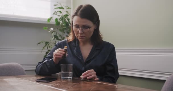 Зрелая женщина капает капли со стаканом воды, пьет успокоительное, дома в пижаме — стоковое видео