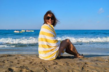 Olgun bir kadın sahilde oturuyor, omuzlarında plaj havlusuyla güzel bir bayanı dinlendiriyor.