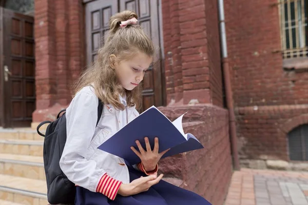 Okul bahçesinde oturan sırt çantalı kız çocuğu defter okuyor, kopya alanı — Stok fotoğraf