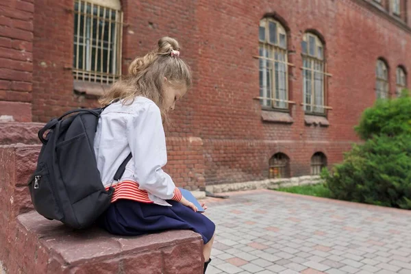 Girl child with backpack sitting in school yard reading notebook, copy space — Stock Photo, Image