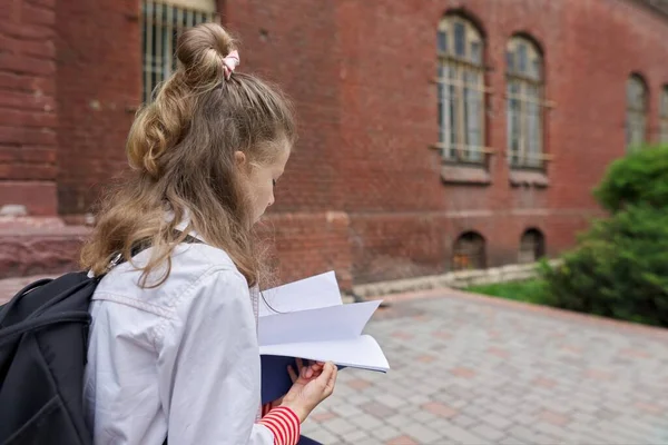 Дівчинка з рюкзаком, що сидить у шкільному дворі, читає блокнот, копіює простір — стокове фото