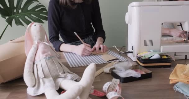 Adolescente menina costura brinquedo coelho boneca sentado em casa, pintura boneca detalhes sobre tecido, padrões de papel — Vídeo de Stock