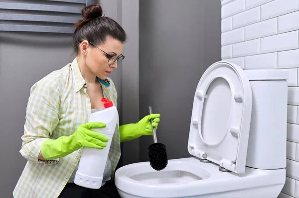 Домохозяйка моет и дезинфицирует туалет, в перчатках с моющим средством и щеткой — стоковое фото