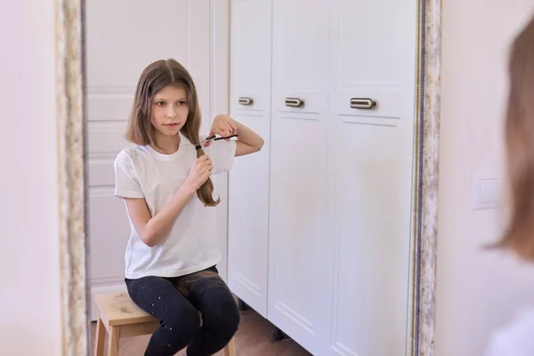 Смішна дівчинка, яка ріже волосся ножицями, сидить вдома перед дзеркалом — стокове фото