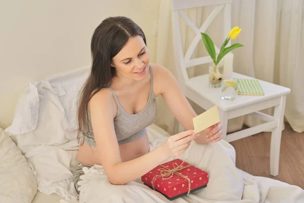 Молода красива вагітна жінка з подарунковою коробкою, в ліжку читає листівку — стокове фото