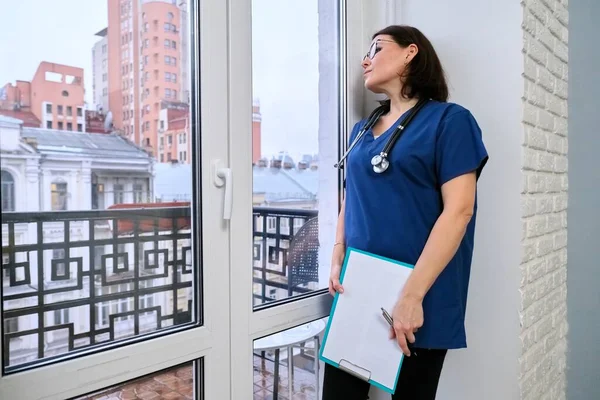 Жіноча медсестра з медичним записом стоїть в офісі, дивлячись у вікно — стокове фото
