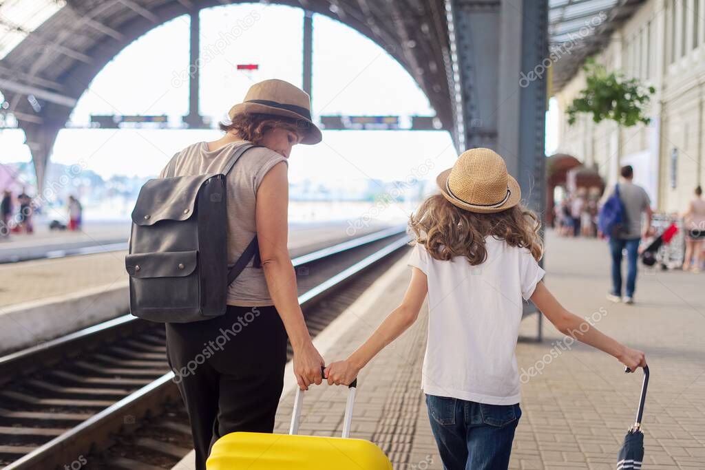 Happy mother and daughter child walking together at railway station with suitcase, back view