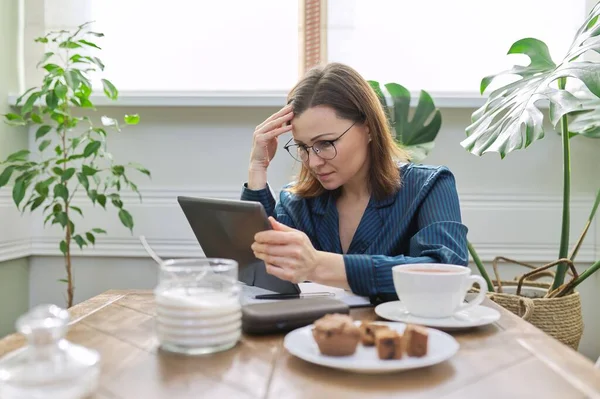Серйозна зріла жінка працює вдома, використовуючи цифровий планшет, бізнес-ноутбук — стокове фото