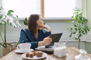 Evde çalışan ciddi olgun bir kadın dijital tablet ve iş defteri kullanıyor.