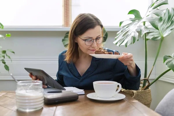 Усміхнена зріла жінка працює вдома, снідає і використовує цифровий планшет, бізнес-ноутбук — стокове фото