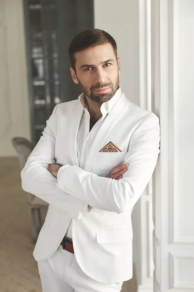 Elegante giovane bell'uomo con la barba in un abito classico bianco. Gli interni degli hotel. Studio fotografico. Divano. Finestra. Sedia. Lampadario — Foto Stock