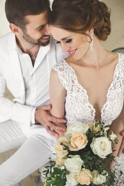 Κομψό γαμήλιο ζευγάρι γαμπρού και της νύφης, θέτοντας σε ένα λευκό στούντιο. Ευτυχές ζεύγος γέλιο. Λευκό φόρεμα. Λευκό κοστούμι. Νεολαία. Γάμου. Πολυέλαιος. Καναπές. Η πόρτα. — Φωτογραφία Αρχείου