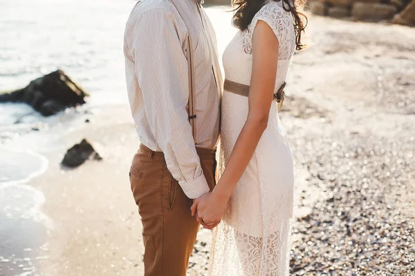 Bruden och brudgummen håller hand på stranden. Solnedgång. — Stockfoto