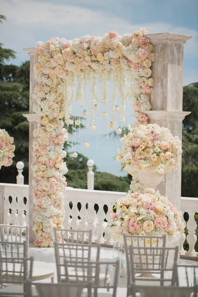 Arco de boda con flores frescas, jarrones, sillas blancas para invitados en el fondo del océano azul . — Foto de Stock