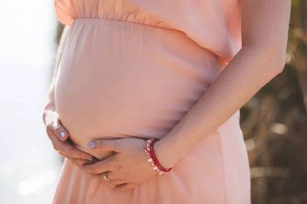 Close-up foto van jonge zwangere vrouw in elegante roze jurk aanraken van haar buik met handen. — Stockfoto