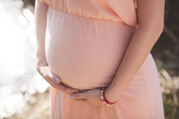 Foto de cerca de la joven embarazada en vestido rosa elegante tocando su vientre con las manos . —  Fotos de Stock