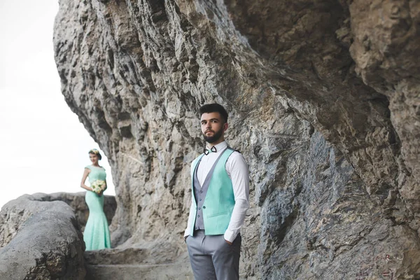 A noiva e o noivo na natureza nas montanhas perto da água. Terno e vestido Tiffany cor. O noivo olha para a distância . — Fotografia de Stock