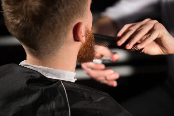 Master taglia capelli e barba nel negozio di barbiere. Il parrucchiere fa l'acconciatura usando forbici e un pettine in metallo . — Foto Stock