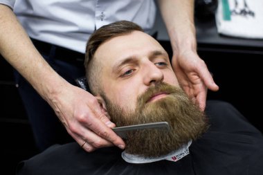 Usta berber dükkanında saç ve sakal kesiyor. Kuaför, makas ve metal tarak kullanarak saç stili yapıyor..