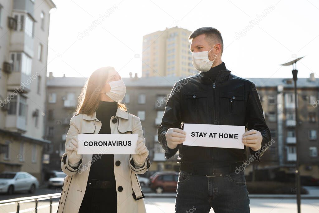 A woman and a man in protective medical masks hold a placard with the words stay at home and coronavirus