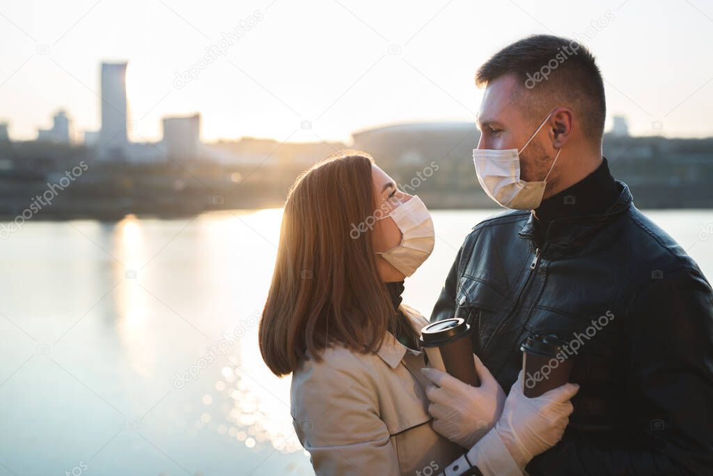 Young couple, a man and a woman in medical masks and gloves, drink coffee from disposable cups on the street and look at each other against the background of the river and the city. Quarantine