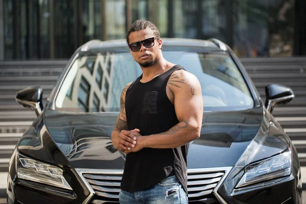 Μαύρος άνδρας με γυαλιά ηλίου στέκεται κοντά στο αυτοκίνητο με το σύγχρονο κτίριο στο παρασκήνιο. — Φωτογραφία Αρχείου