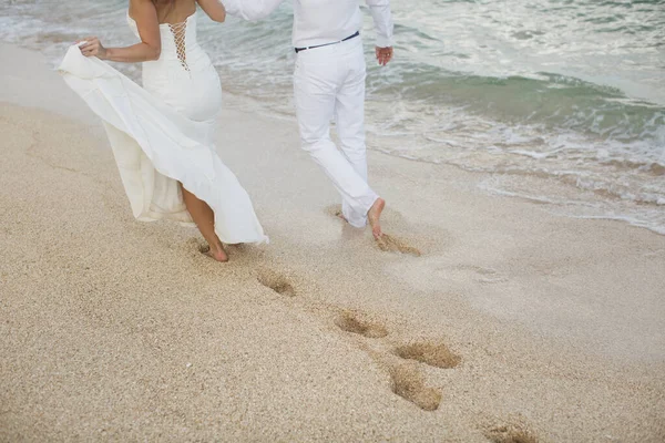 Braut und Bräutigam gehen Hand in Hand in den Sand. Fußabdrücke im Sand in der Nähe des Ozeans. — Stockfoto