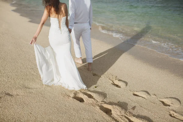 A menyasszony és a vőlegény kéz a kézben sétálnak a homokban. lábnyomok a homokban az óceán közelében. — Stock Fotó