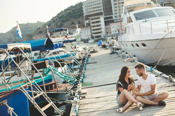 Los amantes, chico y chica, están sentados en un muelle de madera, tomados de la mano y riendo —  Fotos de Stock