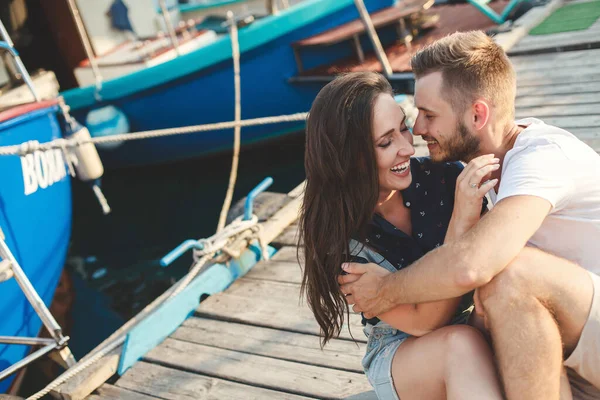 Gli amanti, ragazzo e ragazza, sono seduti su un molo di legno, tenendosi per mano e ridendo — Foto Stock