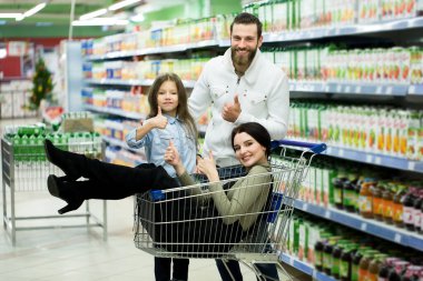 Güzel genç ebeveynler ve onların sevimli küçük kızları süpermarkette yiyecek seçerken gülümsüyorlar. Kızlar orada otururken babam alışveriş arabasını itiyor..