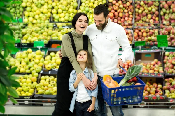 Portret wesołej rodziny stojącej z pełnym wózkiem w supermarkecie w dziale warzyw na tle lady z jabłkami. — Zdjęcie stockowe