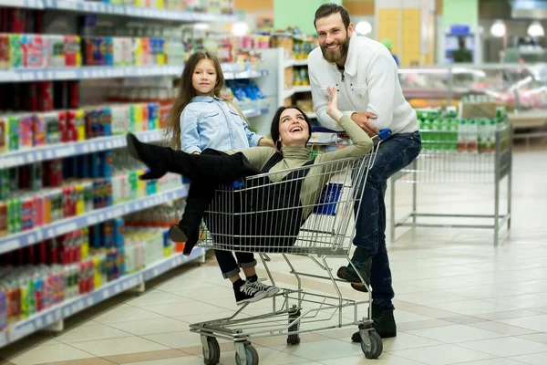 Молодые родители и их симпатичная маленькая дочь улыбаются, выбирая еду в супермаркете. Отец толкает тележку с покупками, пока девочки сидят там . — стоковое фото