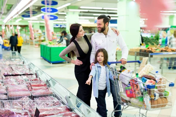 Красивые молодые родители и их милая маленькая дочь улыбаются, выбирая мясо в супермаркете . — стоковое фото