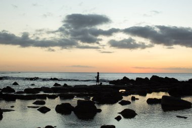 Bir balıkçı okyanustaki siyah taşlarda balık tutuyor..