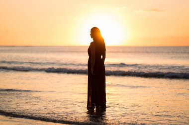 Günbatımı arka planında güzel bir kadın. Denizin üzerinde renkli bir şafak. Kaygısız kadın sahilde günbatımının tadını çıkarıyor. Mutlu yaşam tarzı.